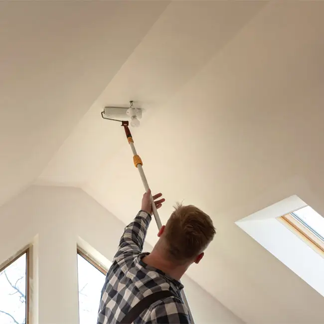 A man stands on a ladder, painting the ceiling of a room with a roller brush.